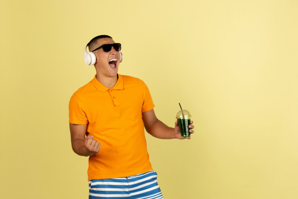 一个年轻的微笑英俊的亚洲人在明亮的夏季衣服与耳机隔离在黄色的工作室背景。图片下载