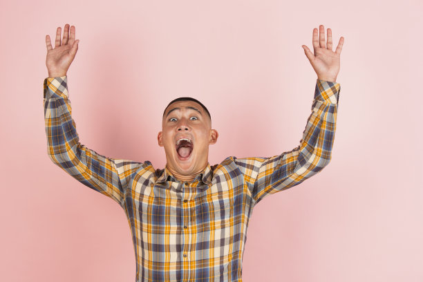 年轻的亚洲人的肖像在格子衬衫与举起的双手孤立在浅粉色工作室背景。图片下载