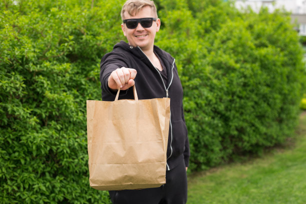 在大自然的绿色背景下，男子手拿外卖生态纸袋。在任何天气条件下24小时送货给客户。图片下载