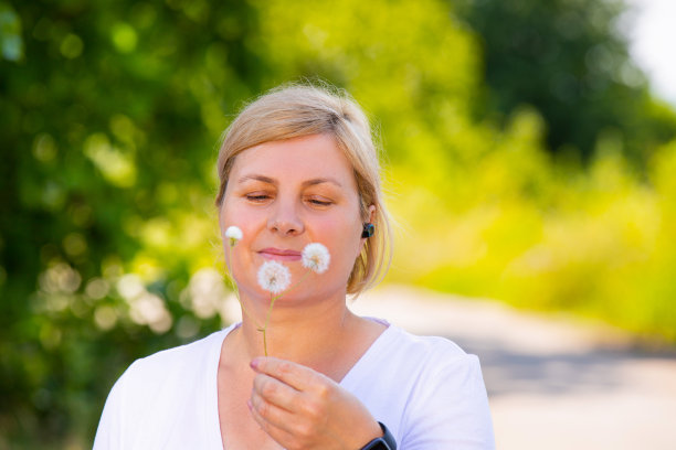 在这张特写照片中，一位漂亮的金发老年女性慢跑时戴着数码手表和耳机，发现了蒲公英花，摘了下来，抱在怀里欣赏这些花。图片下载