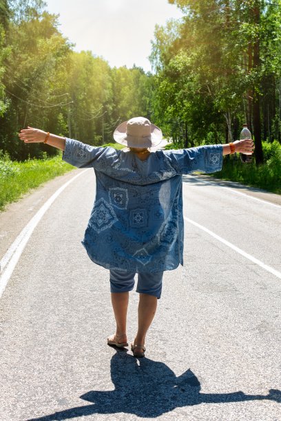 一位上了年纪的妇女一边喝着解渴的饮料，一边在路上走着，享受着阳光明媚的一天。退休老人散步时的积极情绪图片下载