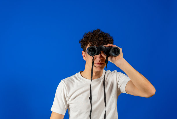 年轻人站在孤立的蓝色背景下，用双筒望远镜望着远方图片下载