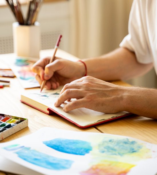 一位年轻的艺术家坐在一张木桌旁，在笔记本上画画。漂亮的男性手拿着彩色铅笔。特写镜头。阳光和宜人的气氛。diy概念。真正的艺术家在行动图片下载
