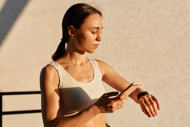 在户外拍摄的照片中，苗条的美女身穿白色运动上衣，深色头发，站在夕阳下，手里拿着智能手机，在训练前系好臂章图片下载