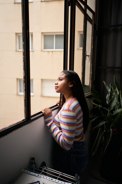 垂直的照片。非裔美国女孩靠近窗户。从家里面带微笑地看着外面。图片下载