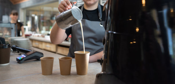 一名女咖啡师将牛奶倒入金属壶里的咖啡杯中。三纸杯咖啡和牛奶。卡布奇诺的制作过程。拿铁咖啡烹饪概念。图片下载