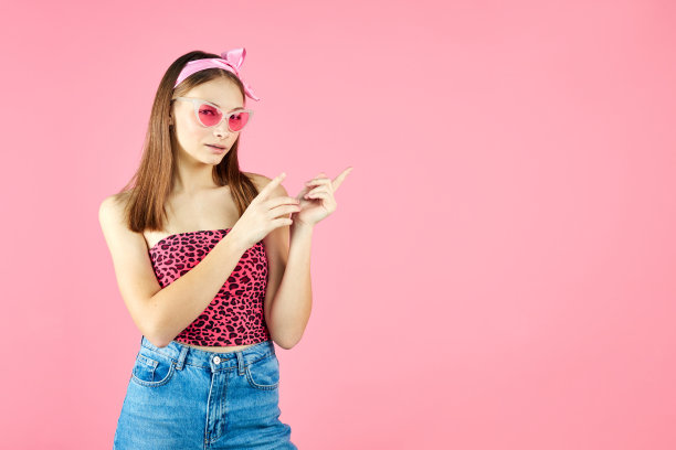 戴着时髦眼镜的酷潮女。正面的年轻女子看着相机在粉红色的背景。白人女大学生摆姿势，用手指。图片下载