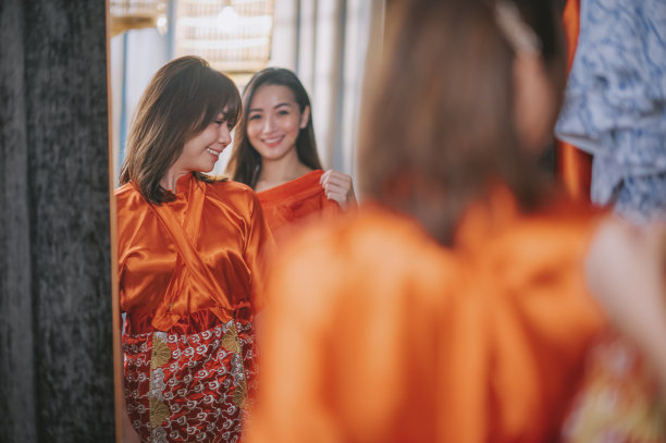 在婚纱店，亚洲中国新娘在姐姐的帮助下，对着镜子试穿中国传统婚纱图片下载