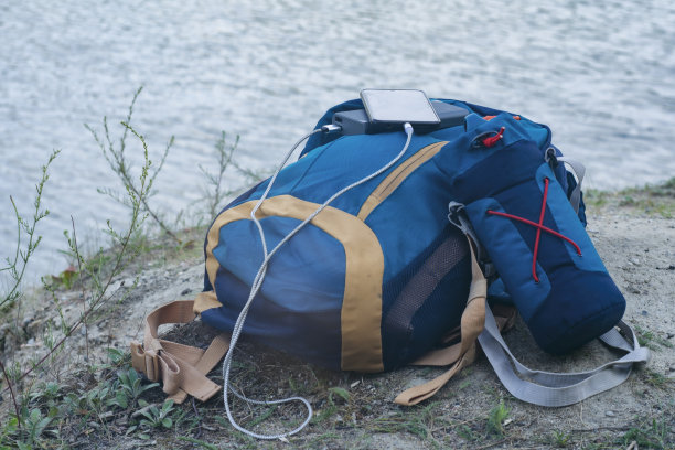 智能手机使用便携式充电器充电。Power Bank在户外为手机充电，并配有旅游背包，以自然和河流为背景。图片下载