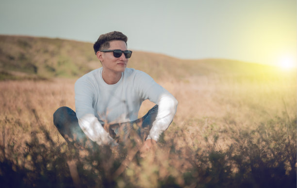 微笑的青年男子坐在草地上，微笑的青年男子戴着眼镜坐在田野上，幸福的概念在田野图片下载