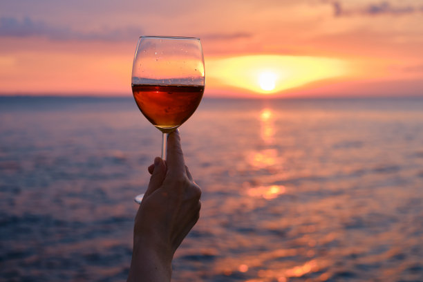 在海边夕阳的余辉中，一个女孩手中的一杯红酒。酒精饮料品尝。美丽的日落和海滩上的美酒。浪漫的夜晚开胃酒。玻璃特写镜头图片下载