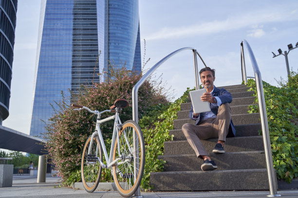 一位商人坐在办公楼前的楼梯上，在电子平板电脑上阅读电子邮件。一个人骑着自行车穿过城市。图片下载