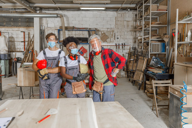 一群戴着防护面具的多文化木匠在一家木工工厂对着镜头微笑。图片下载