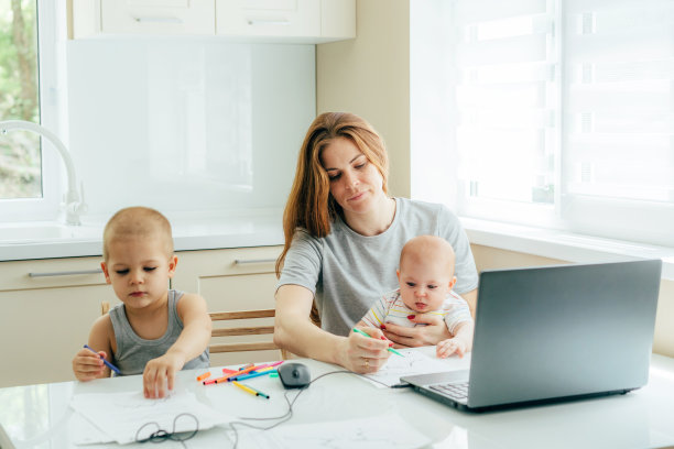 年轻疲惫的母亲一边用笔记本电脑工作，一边在厨房的餐桌旁照顾孩子。图片下载