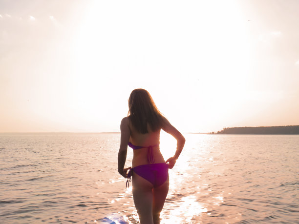 年轻迷人的女子穿着比基尼泳装，在橙色的阳光下，水的背景图片下载