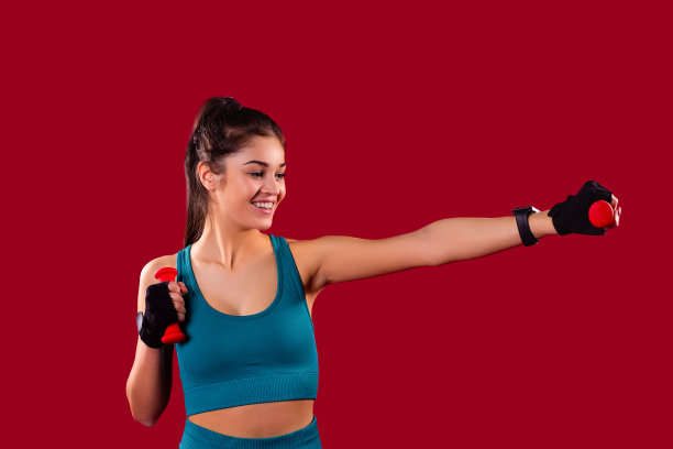 一个美丽的运动年轻女子的肖像站着，手里拿着哑铃，左臂向前伸展。图片下载