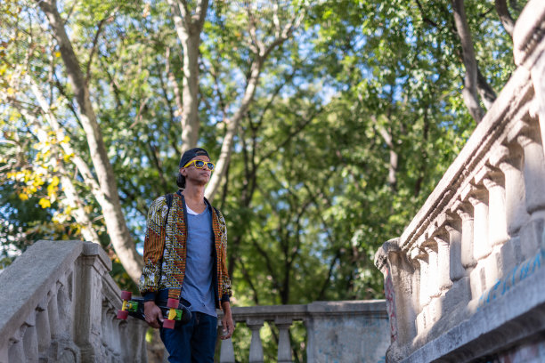 西班牙潮人与迷你溜冰在一个姿势，拉丁美洲男子与时髦和时尚的风格在城市公园，非洲式男子溜冰者在放松的一刻图片下载