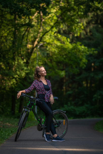 一个夏天，一个年轻的女人穿着衬衫和打底裤，骑着自行车站在公园的柏油路上，看着旁边的某个地方。垂直图图片下载
