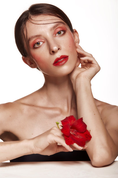 漂亮的女人手里拿着红花靠近脸裸露的肩膀清楚的皮肤模特用粉红色的妆。皮肤护理和水疗概念在白色的背景。看相机的女人图片下载