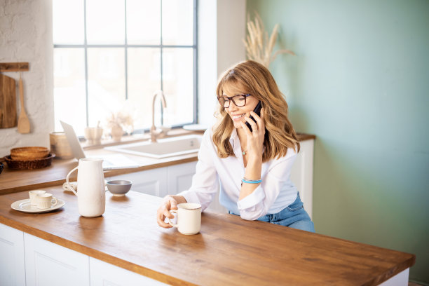 快乐的女人一边在厨房喝早茶一边打电话图片下载