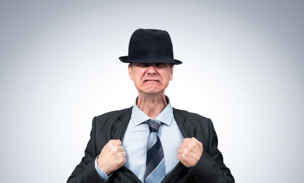 情绪紧张的愤怒男子身穿西装，戴着帽子，在浅灰色背景下用手握住他的夹克。文件包含了隔离人类的途径。图片下载