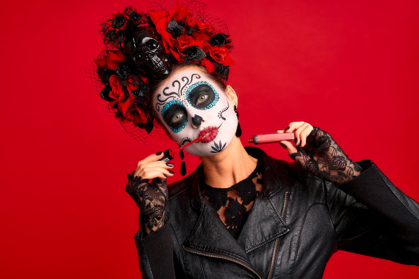 女孩用糖骷髅妆，头上和头盖骨上戴着花环，戴着蕾丝手套，穿着皮夹克，在红色背景下测试新的唇彩图片下载