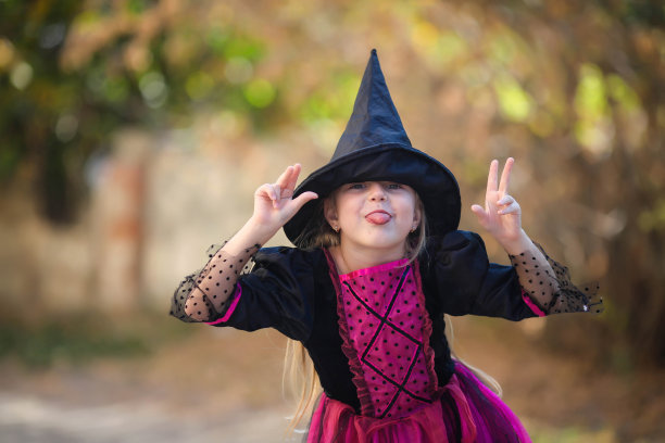 一个穿着女巫服装的小女孩的肖像，躲在黑色的帽子后面，露出她的舌头。万圣节的节日。本空间图片下载