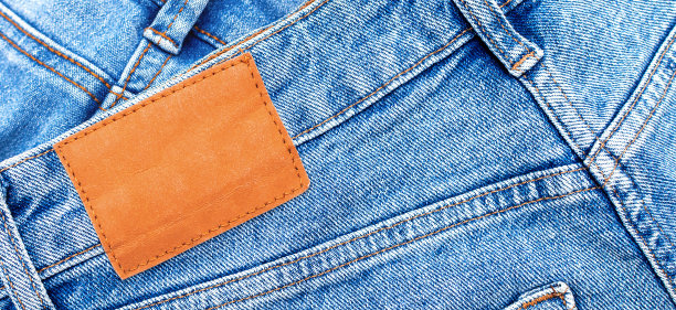 牛仔面料，蓝色牛仔裤和空白皮革标签，舒适的休闲裤和衣服的特写。棉织物表面有接缝和橙色线缝。带有复制空间的横幅背景图片下载