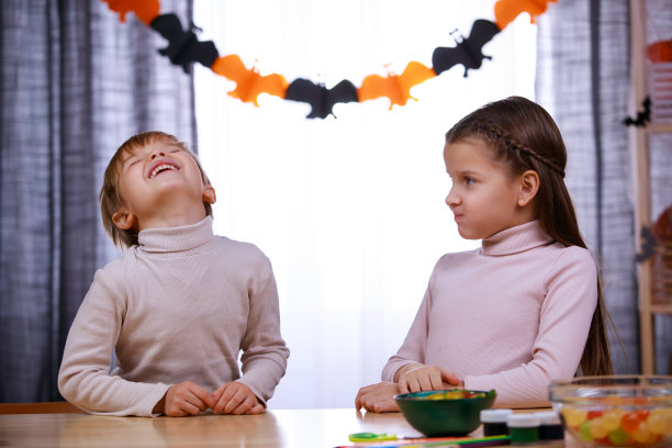 男孩和女孩坐在一张摆满糖果和南瓜的桌子旁，房间里装饰着万圣节。妹妹嚼着糖果，弟弟则对着它狂笑。快乐的家庭聚会在家里。近距离图片下载