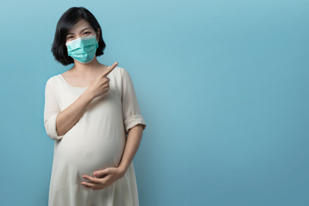 微笑的脸，快乐的亚洲孕妇戴着医用防护口罩在蓝色隔离背景指向复制空间。图片下载