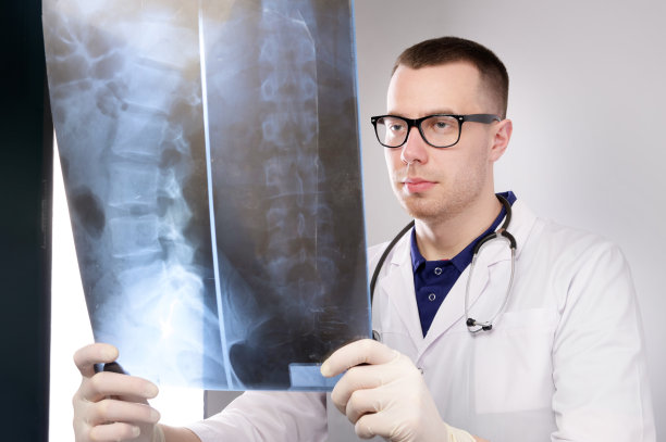 年轻的放射科医生戴着眼镜，穿着医疗服，看着灰色背景下的脊柱腔x射线照片。图片下载