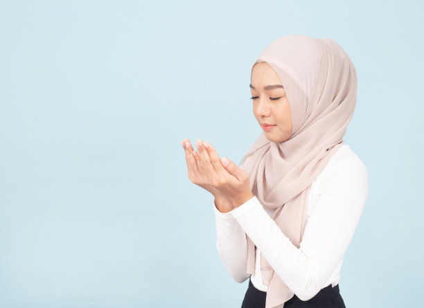 隔离在蓝色背景上。微笑的亚洲年轻穆斯林妇女的肖像在希贾布头巾祈祷。图片下载