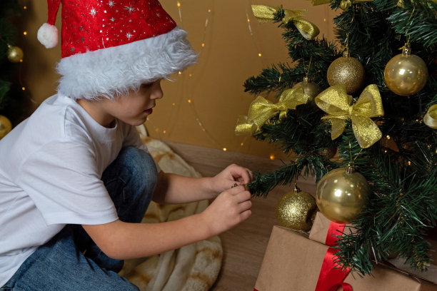 一个戴着新年帽的男孩用金球装饰圣诞树。有选择性的重点。图片下载