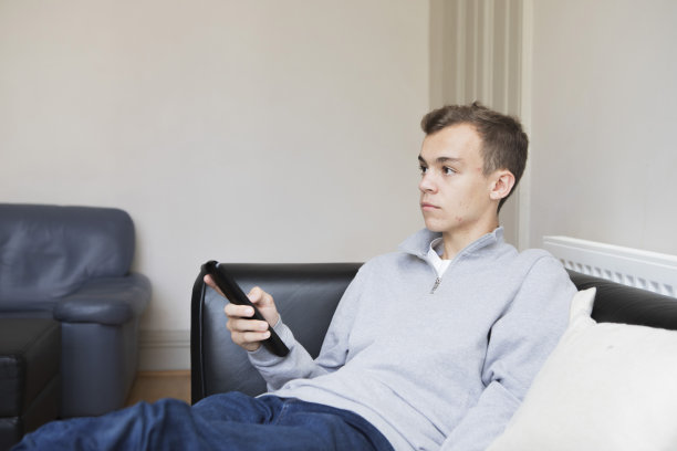 一名男大学生跷着脚坐在黑色沙发上，拿着遥控器看电视图片下载