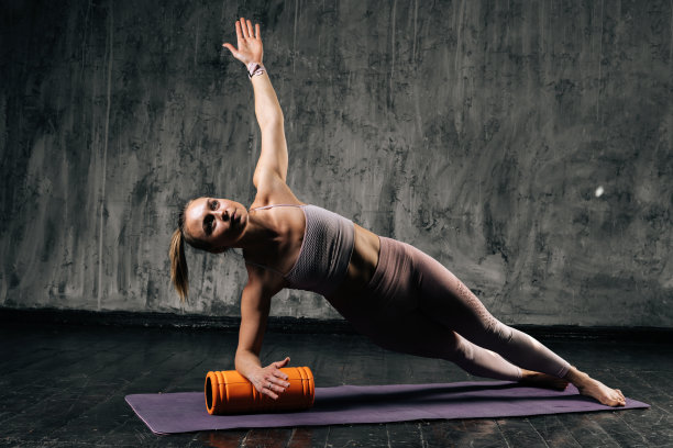 肌肉发达的年轻运动女性与完美美丽的身体穿着运动服做侧板姿势使用泡沫滚轮按摩器。图片下载