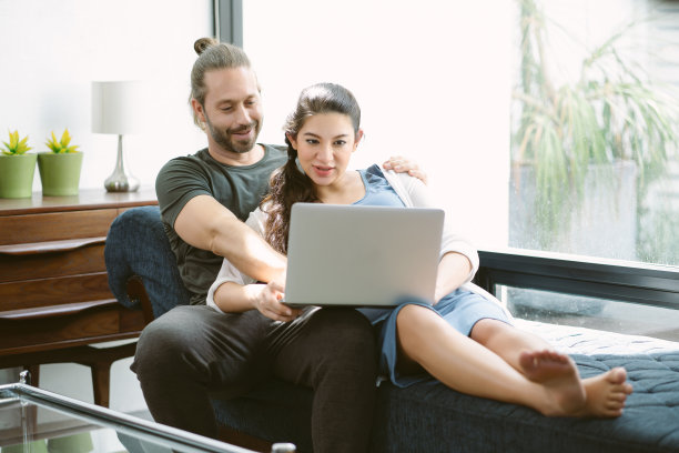 这是一对可爱的怀孕夫妇坐在沙发上用笔记本电脑共度时光的照片。应用程序用户，购物者为孩子选择商品的概念。图片下载