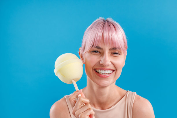 一名粉红色头发的快乐女子对着镜头微笑，手里拿着一个大棒棒糖冰淇淋，在蓝色背景下摆出孤立的姿势图片下载