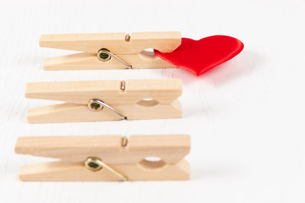 白色木桌背景上，三个带有金属弹簧的木衣夹夹着一颗红心。特写镜头图片下载