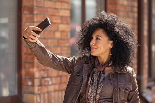时尚的年轻非洲裔美国女人的肖像在手机自拍。黑发卷发，身穿棕色皮夹克在街道上摆姿势，背景是模糊的砖建筑。近距离图片下载