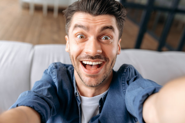 一名穿着便服的白人青年坐在沙发上自拍，一边拍照一边做鬼脸，手里拿着智能手机，热情地看着相机，微笑着图片下载