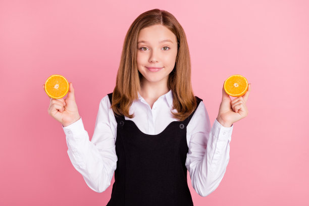 漂亮甜美的女学生穿着黑白制服，拿着两片橙片微笑着孤立的粉红色背景图片下载