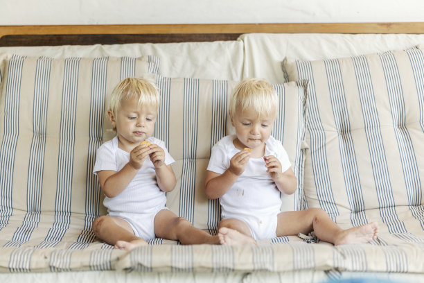 两个漂亮的宝宝在吃饼干。一对金发双胞胎在客厅的沙发上吃饼干。有趣的婴儿照片，一模一样的孩子在一起吃糖果图片下载