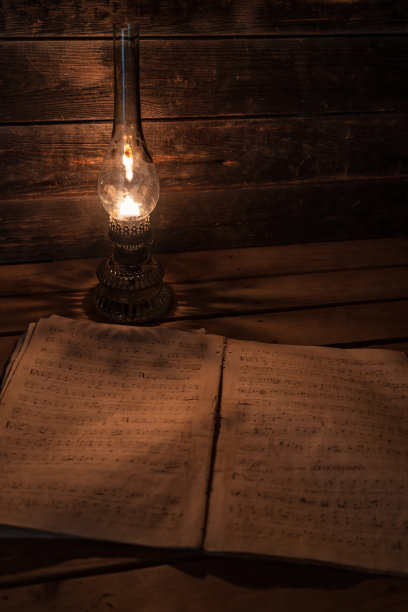 暮霭中，煤油灯的灯光照亮了一本有音符的旧音乐书。有一面古老的木墙和一张桌子。背景。纹理。图片下载