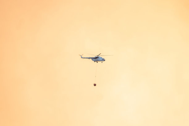 空中消防在黄色多云的天空背景中带着水篮的飞行直升机。图片下载