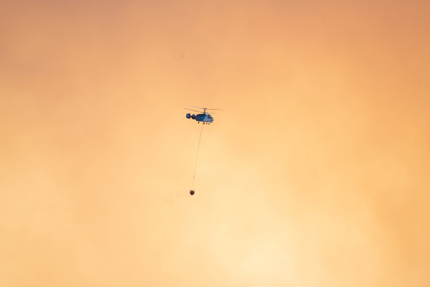 空中消防在黄色多云的天空背景中带着水篮的飞行直升机。图片下载