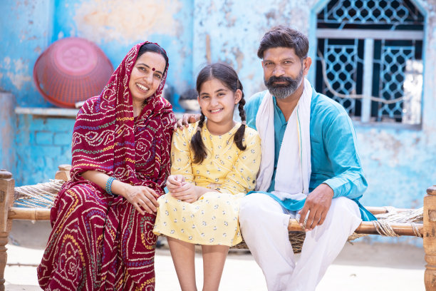可爱可爱的小女儿抱着她的爸爸妈妈，坐在传统的床上，看着镜头，幸福的印度农村家庭。印度父母带着孩子微笑图片下载