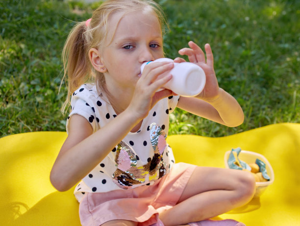 快乐微笑的女孩喝酸奶或牛奶在户外夏季野餐。手里拿着白色的塑料瓶，脸上还沾着酸奶图片下载
