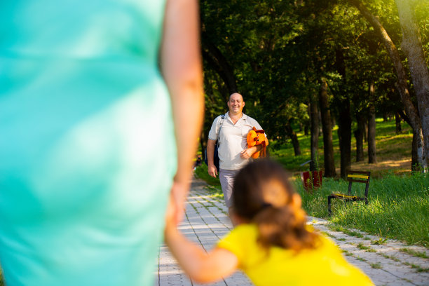 妈妈和小女孩的后视图照片等不及她的爸爸一起在公园度过洞日，他准备迎接他们，手里拿着一个柔软的狗玩具给他的女儿。图片下载