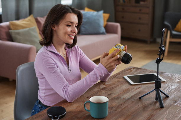 积极有吸引力的年轻白人摄影师坐在桌子上，用三脚架上的智能手机，在准备现场拍摄的时候浏览相机照片图片下载