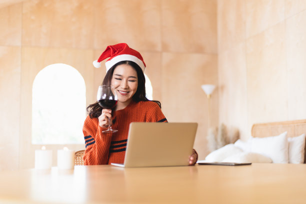 年轻的亚洲女人戴着圣诞帽喝着红酒。美丽的女性与朋友和家人在虚拟视频电话庆祝新年晚会在家里的电脑上。图片下载